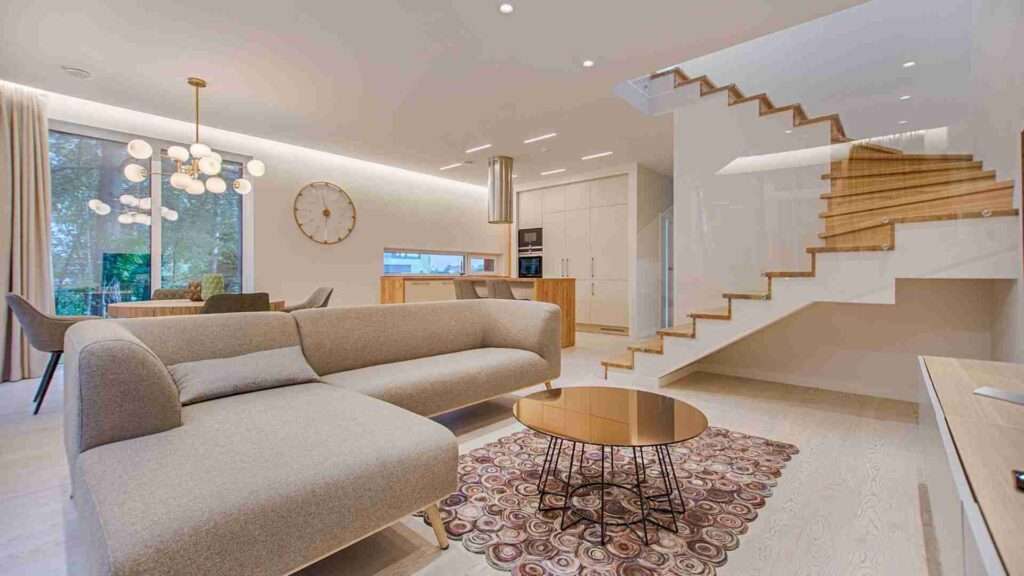 Modernes Wohnzimmer mit beigem Sektionssofa, rundem Couchtisch aus Glas auf gemustertem Teppich, offenem Essbereich, Holztreppe mit Glasgeländer und großen Fenstern, die viel Tageslicht hereinlassen.